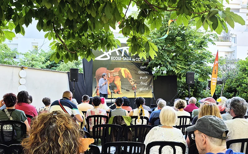 Le public, assis dans le jardin sous les arbres, écoute un intervenant au micro devant la scène, pour les vingt ans du GEM Lesseps