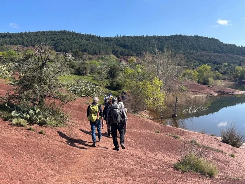 Les randonneurs d’Espoir Hérault marchent en file sur la terre rouge, au bord du lac du Salagou